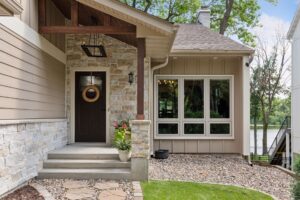 Welcoming lakeside entry with stone veneer, dark wood gable, tan siding, and large grid windows.