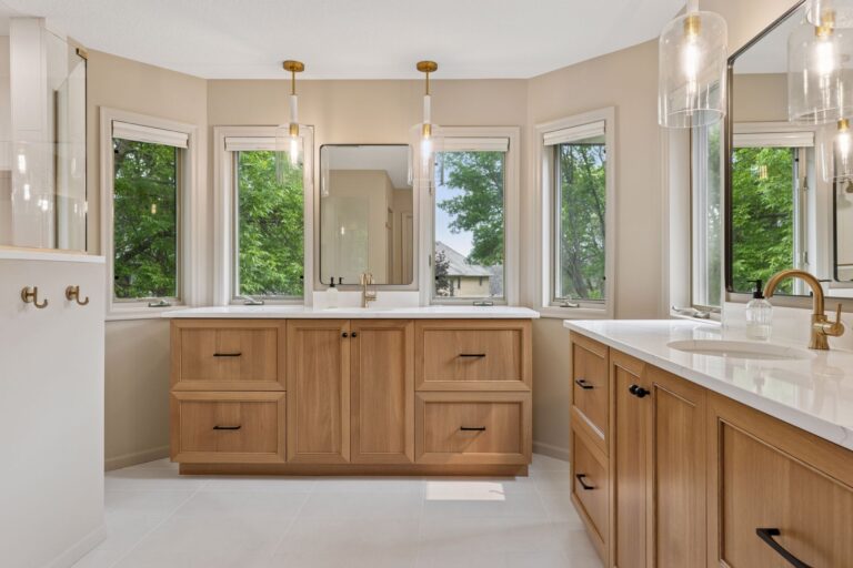 Modern bathroom with natural wood cabinets, white countertops, gold fixtures, and large windows providing ample natural light.