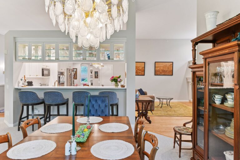 Open-concept dining and kitchen area with glass-front cabinets, blue barstools, a statement chandelier, and traditional wood furniture.